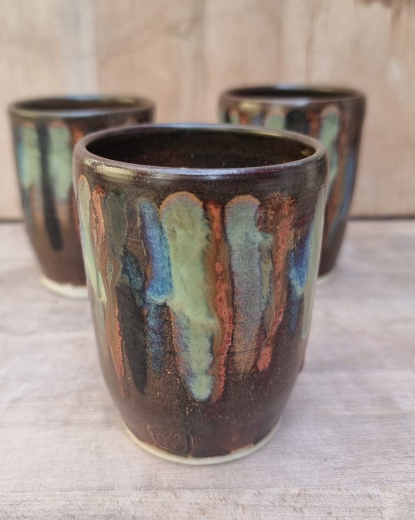 brown and coloured beakers. one in foreground with two behind. wooden benchand backing.