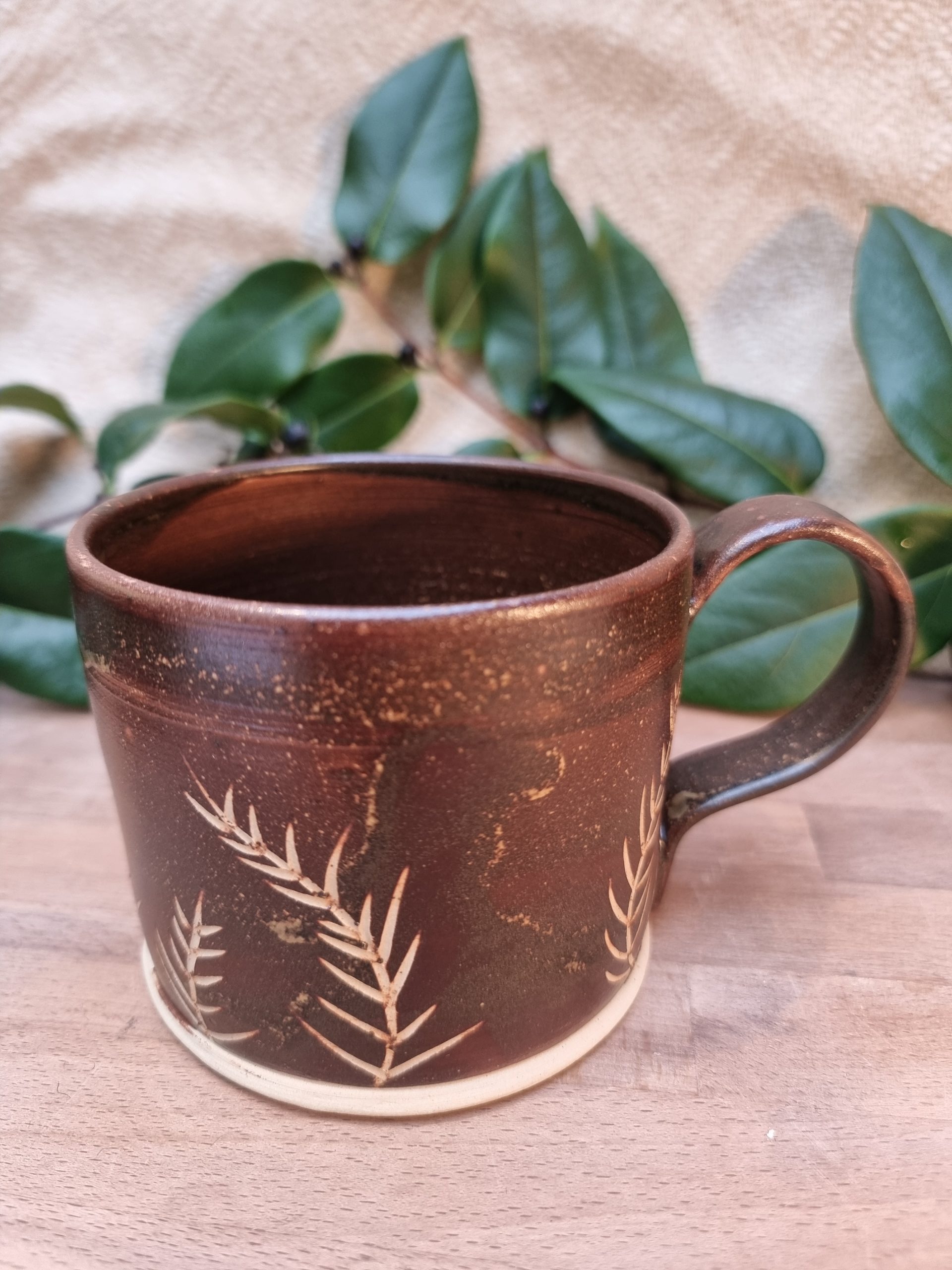Brown mug with white yew leaf patterns. Over a wooden table and greenery.