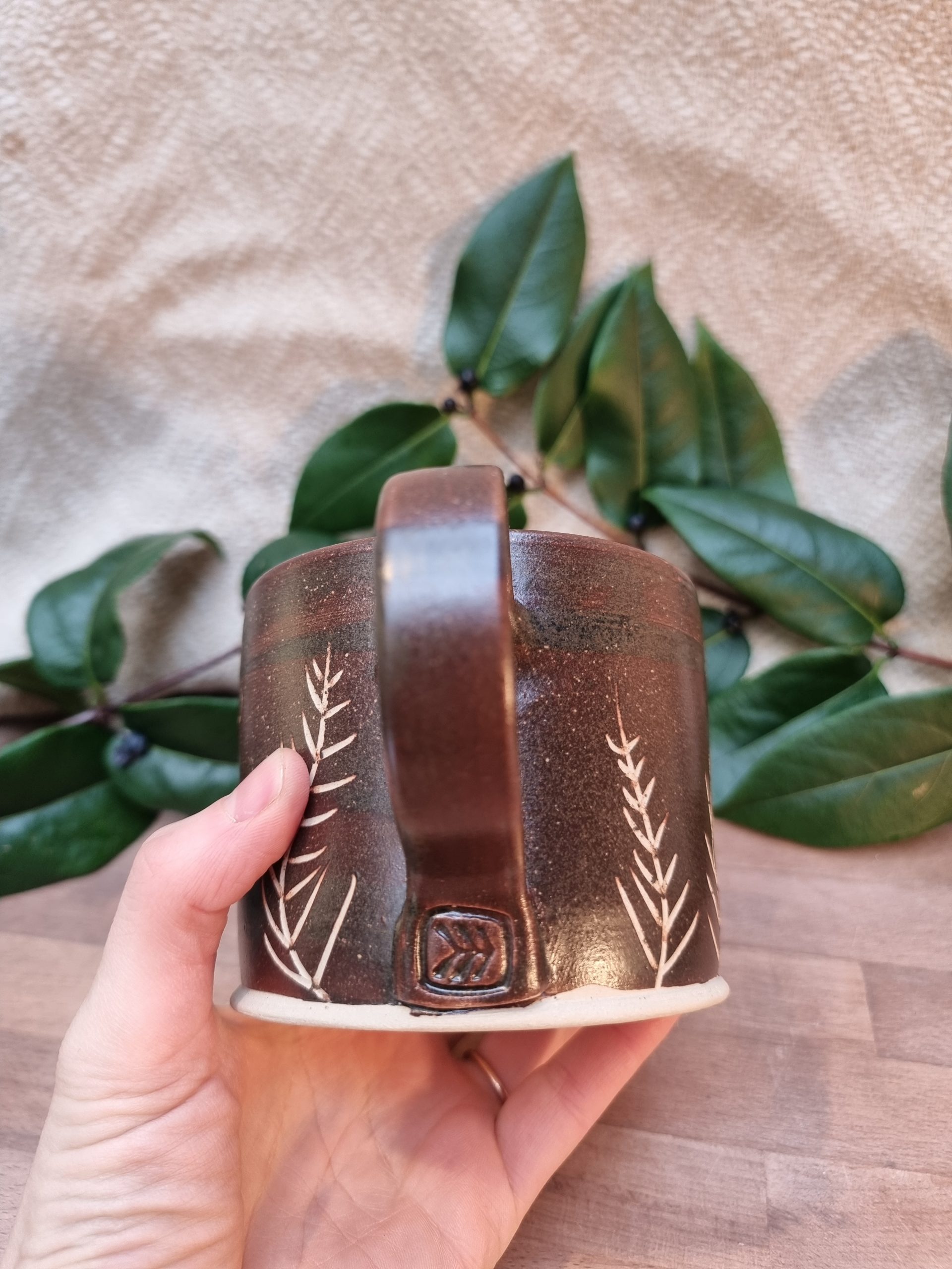 Brown mug with white yew leaf patterns. held in a hand over a wooden table and greenery.