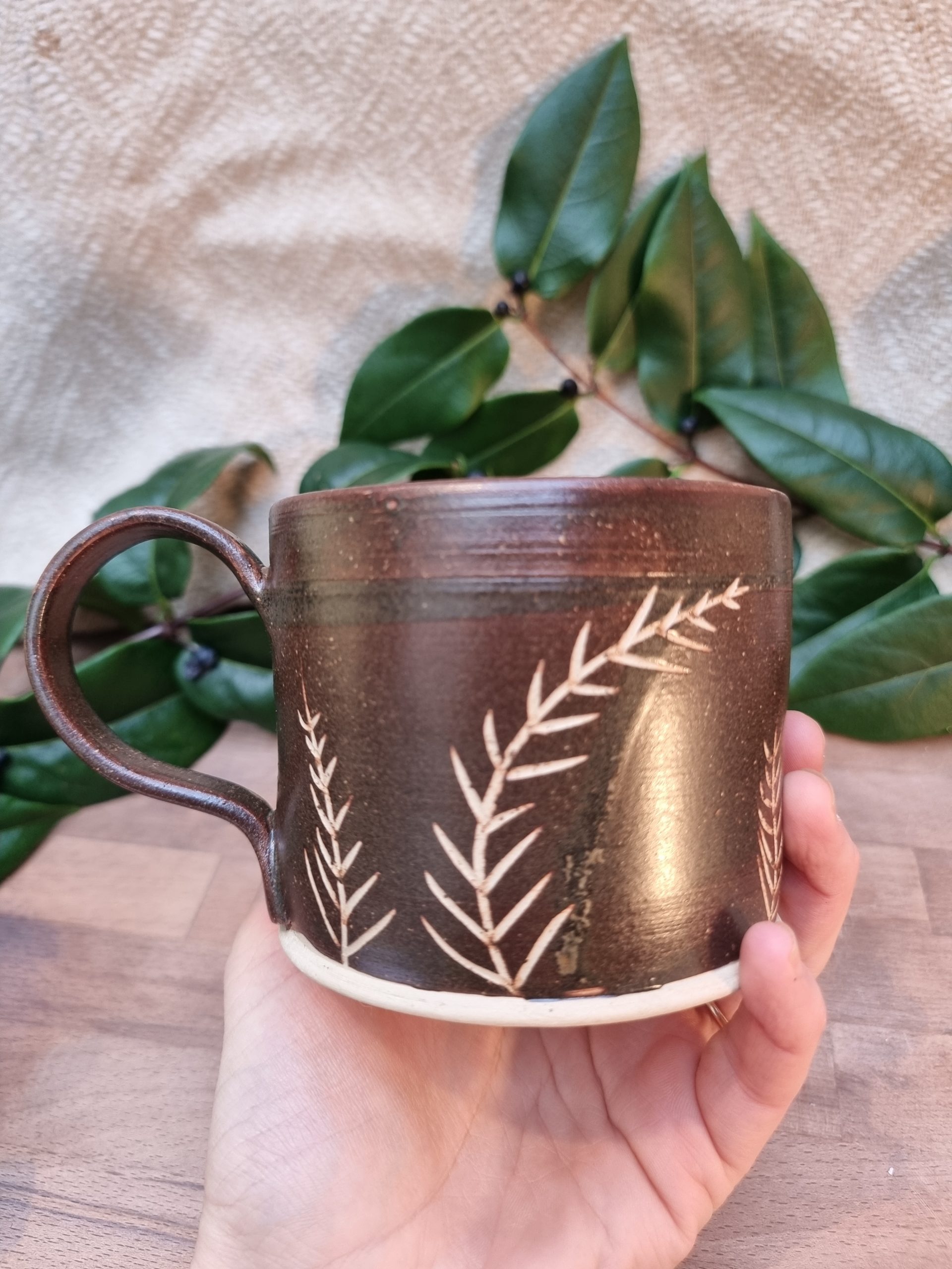 Brown mug with white yew leaf patterns. held in a hand over a wooden table and greenery.